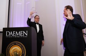 A man dressed in a black tux, standing behind a podium, holding a clear plaque in the air and another man dressed in a suit walking up to accept the award. 