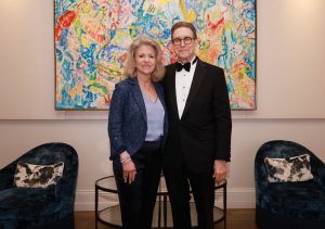 A woman dressed in a formal pant suit and a man dressed in a black tux standing together in front of a colorful painting