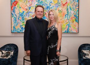 Man dressed in black suit and black bow tie standing with a woman dressed in a black dress in front of a colorful painting