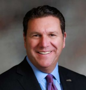 Man with short dark hair wearing a light blue dress shirt, red and blue tie and dark suit jacket