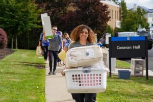 Woman carrying a laundry basket filled with items and people walking behind her carrying items into a dorm.