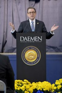 Man wearing a dark suit, white dress shirt and blue tie, standing behind a podium speaking to an audience