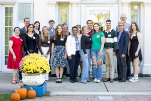 Group of men and women standing on the patio with a white house behind them and pumpkins and yellow mum plants as accents