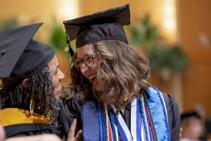 Two women wearing commencement caps and gowns smiling at each other on stage.
