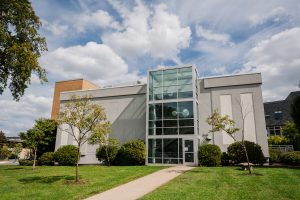 Large gray building with three-story glass entrance. Lawn and trees in front of the building