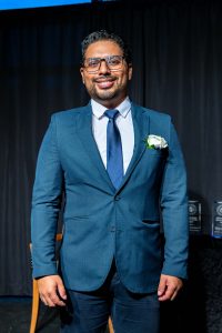 Man dressed in a blue suit with a white shirt and blue tie standing in front of a dark background.