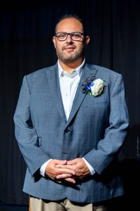 Man wearing glasses, tan dress pants, a white dress shirt and a blue sport coat standing in front of a dark curtain.