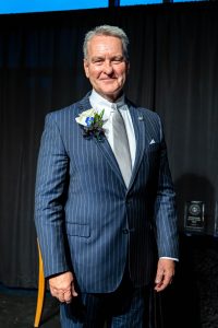 Man dressed in a blue pinstripe suit with a white shirt and gray tie standing in front of a dark background.