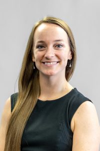 Woman with long light brown hair wearing a sleeveless black shirt smiling in front of a gray background