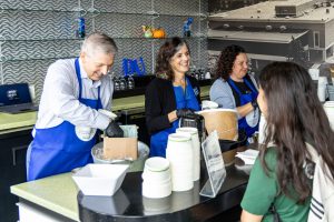 Two women and a man wearing blue aprons, scooping ice cream behind a counter and serving it to students