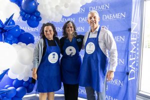 Two women and a man wearing blue aprons, standing arm-in-arm in front of a blue Daemen backdrop with blue and white balloons.