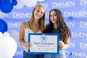 Two young female students holding an oversized Dean's List Recipient certificate standing in front of a blue Daemen backdrop with blue and white balloons.