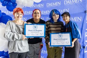 Four young female students holding two oversized Dean's List Recipient certificates standing in front of a blue Daemen backdrop with blue and white balloons.