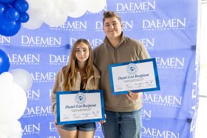 Young female and male student holding an oversized Dean's List Recipient certificate standing in front of a blue Daemen backdrop with blue and white balloons.