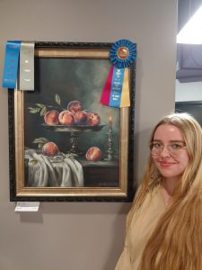 Young woman with long blonde hair and glasses standing next to framed painting of peaches on a platter with three ribbons: One blue, one gray, and one with blue, yellow, and red 