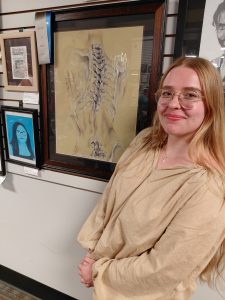 Young woman with long blonde hair and glasses, standing next to a framed figure drawing of a skeletal figure of a spine with blue and gray ribbons on it
