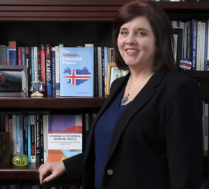 Woman with dark brown/reddish shoulder-length hair standing in front of a book case with several books on the shelves