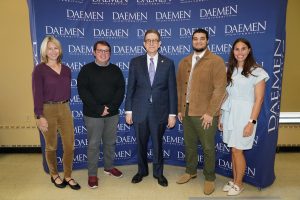 Group of 3 men and 2 women dressed nicely standing in front of a blue Daemen backdrop