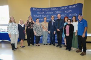 Eight women and three men dressed nicely standing in front of a blue Daemen backdrop