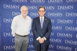Two men dressed nicely standing in front of a blue Daemen backdrop