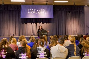 Woman standing behind a podium with a blue Daemen banner hanging behind her, speaking to a seated crowd 