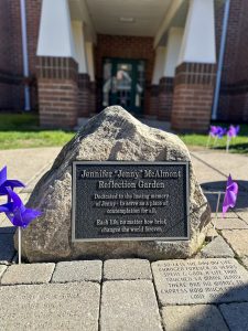 Memorial stone with text on it and purple pinwheels on either side of it with a building behind it and more purple pinwheels in the grass