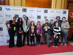 10 young male and female students standing on a red carpet in front of a backdrop with several logos