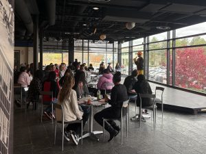 A room filled with tables and chair with people seated at them and windows behind them.