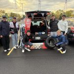 Six young men dressed in pants and hoodies or jackets standing around a trunk of a car that's NASCAR themed.