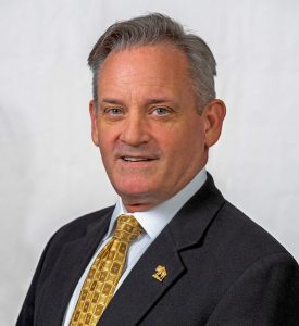 Middle aged male with salt-and-pepper hair wearing a dark suit jacket, white shirt and yellow and gold tie