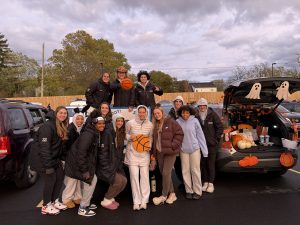 Thirteen young females dressed in pants and jackets, standing in front of cars in a parking lot