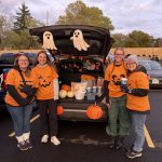 Four women dressed in orange pumpkin shirts standing in front of an open trunk with ghosts, candy and other treats inside