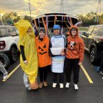 Four young women dressed in colorful halloween costumes standing in front of an open trunk with orange and purple streamers hanging on it.