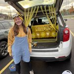 Young female with long blonde hair dressed up as a minion standing in front of a trunk decorated with yello streamers and balloons