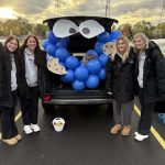 Four young women wearing dark pants and jackets standing in front of the trunk of a car that's decorated as Cookie Monster