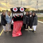 Five young women wearing long pants and jackets, standing by the trunk of a car that's decorated as a red monster