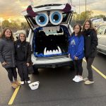 Four women wearing dark pants and jackets standing by the trunk of a car decorated as a monster