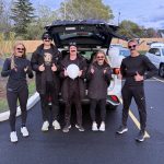 Five young men and women dressed in black pants and jackets, wearing fake mustaches, standing in front of a car