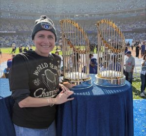 Woman, smiling and wearing a winter hat standing next to two trophies on a baseball field