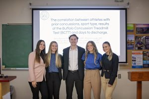 One male and four female physical therapy students standing in front of a classroom with a large presentation screen behind them