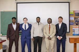 Five male PT students standing in front of a classroom with a large presentation screen behind them