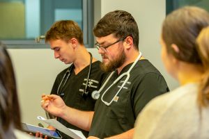 Two male college students wearing scrubs and stethoscopes around their necks, reviewing documents