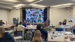 Four adults sitting behind a long table with a presentation displayed on a large screen behind them. Other participants are sitting at tables facing them.