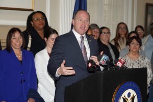 Man dressed in a gray suit, white shirt and tie standing behind a podium speaking with a group of people standing behind him