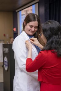 Female graduate nursing student wearing white coat with woman in a red dress putting a pin on her lapel