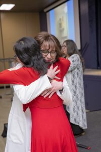 Female graduate nursing student wearing white coat hugging female wearing a red dress in front of a room 