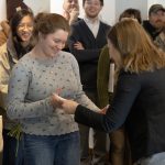 Female professor handing a female art student a white envelope while a crowd looks on