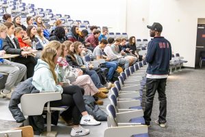 Man wearing a USA jacket standing in the front of an auditorium-style classroom speaking to several students who are seated.
