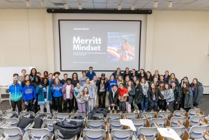 A large group of men and women standing together in the front of an auditorium-style classroom with a large screen with a presentation displayed behind them.