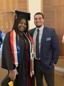 Man wearing a blue suit, white shirt and tie standing next to a woman wearing a graduation cap and gown.